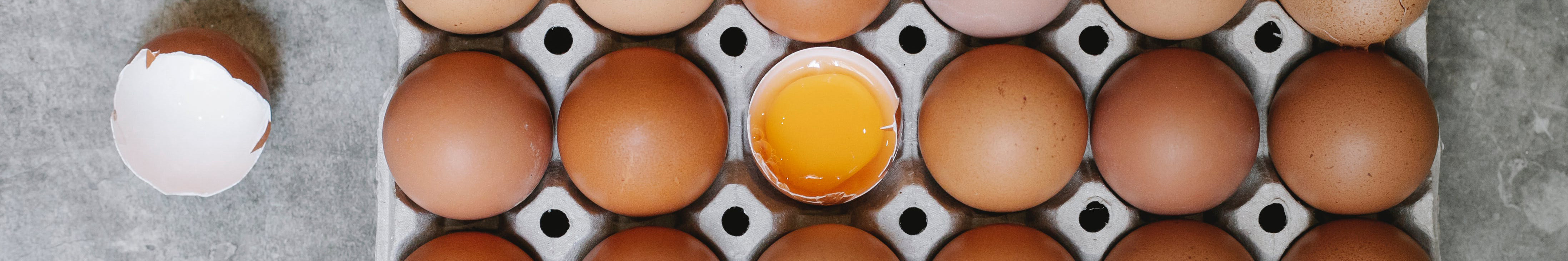 An open carton of eggs, one is cracked in half with the full yolk visible.